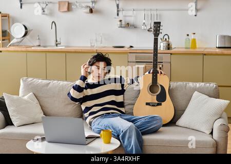 Handsome young Asian man sits comfortably on a couch while practicing guitar, focused and inspired. Stock Photo
