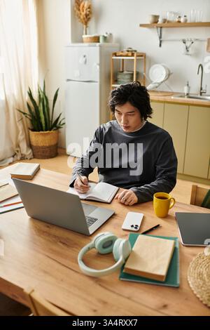 Focused student writes notes while studying on laptop at home during the day in cozy kitchen. Stock Photo