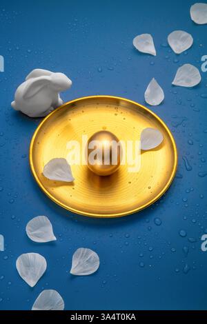 Golden easter egg displayed on a golden plate with a white ceramic bunny and delicate white petals on a vibrant blue background, creating a luxurious Stock Photo