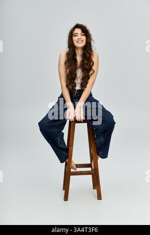 A charming young woman flaunts her fashion as she playfully sits on a wooden stool. Stock Photo
