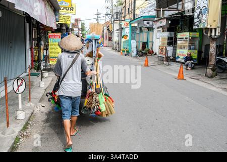 Street vendor wearing traditional hat pushing a cart of cleaning supplies along a quiet street in yogyakarta, indonesia Stock Photo