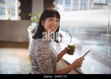Smiling woman enjoying green smoothie while browsing tablet in cozy cafe Stock Photo