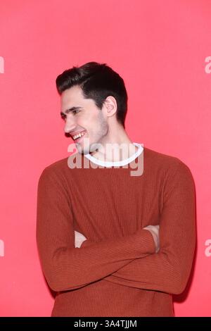 A young man in a brown sweater with arms crossed, posing confidently against a vibrant red background. Stock Photo