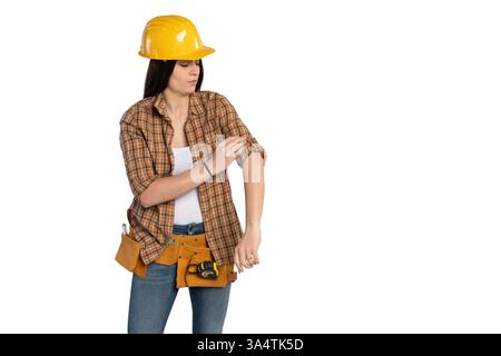 Young female construction worker in a yellow hard hat and plaid shirt rolling up her sleeves, preparing for work. She wears a tool belt with tools, je Stock Photo