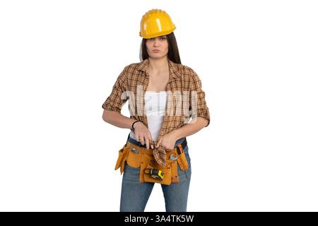 A female construction worker in a yellow hard hat and plaid shirt adjusting her tool belt. She wears jeans and a white tank top, standing confidently. Stock Photo