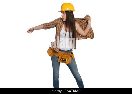Young female construction worker in a yellow hard hat adjusting her plaid shirt. She wears a tool belt with various tools, jeans, and a white tank top Stock Photo