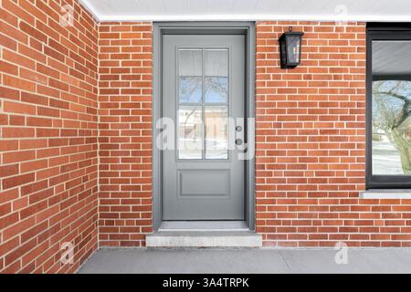 A home's grey front door with frosted glass surrounded by red brick, white ceiling, concrete patio, and black framed window. Stock Photo