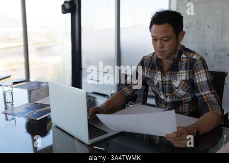 Asian man reviewing documents while working on laptop in modern office, copy space Stock Photo