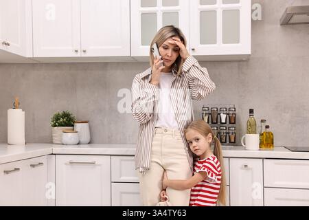 Work at home. Overwhelmed single mother talking by smartphone and her daughter in kitchen Stock Photo