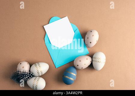 Easter-themed greeting composition with a blank white card in a blue envelope and decorative eggs with golden patterns on a brown background. Top view Stock Photo