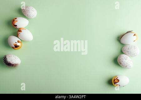 White and gold Easter eggs with speckled patterns and feathers arranged on a pastel green background. Top view, flat lay, copy space. Stock Photo
