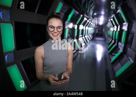 Smiling woman using smartphone in futuristic office corridor with green lights, copy space Stock Photo