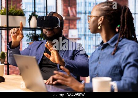 African American man interacts with VR while his girlfriend manages the 3D animation on a laptop at desk. Young couple integrates artificial intelligence and creativity in their workspace. Stock Photo