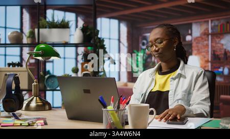 Upset woman receiving bad news from text message on smartphone while using notebook in home office. Sad remote worker troubled by SMS received on mobile phone while using laptop, camera A Stock Photo