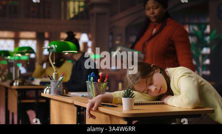 Teacher waking up her student sleeping on the study desk at the library, hosting private lessons with the tired overstimulated schoolgirl. Professor mentoring her fatigued pupil. Camera B. Stock Photo
