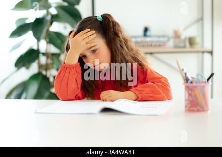 Tired bored caucasian schoolgirl in a red blouse sits at a white desk, resting her forehead on her hand while looking at her workbook with a frustrated expression during doing homework at home Stock Photo