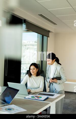 In a bright office, two women engage in a collaborative discussion while reviewing data on a laptop, showcasing teamwork and professionalism on a prod Stock Photo