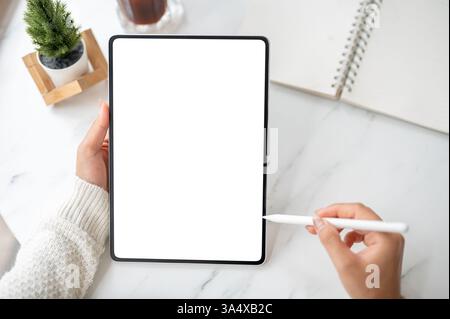 Close up of the left hand holding blank screen or white screen tablet and stylus pencil pointing at the screen in right hand, on the marble table. Stu Stock Photo