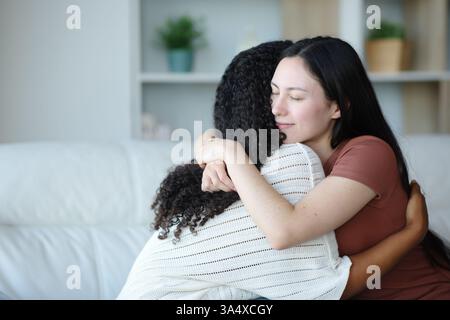 Satisfied asian woman hugging a black friend sitting on a couch at home Stock Photo