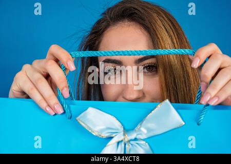Young woman with long brown hair holding light blue shopping bag with ribbon in front of face on blue background Stock Photo