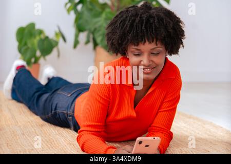 Happy African American woman using smartphone while lying on the floor at home. Stock Photo
