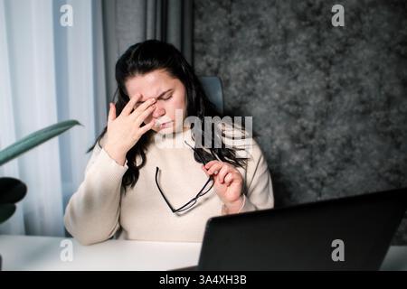 Tired woman working at home with laptop holding eyeglasses rubbing eyes and feeling fatigue. Stress and frustration concept. Tired eyes. Stock Photo