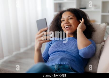 Young African American woman lying on couch, laughing while looking at smartphone Stock Photo