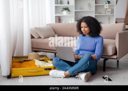 Young African American woman using laptop at home while packing for trip Stock Photo