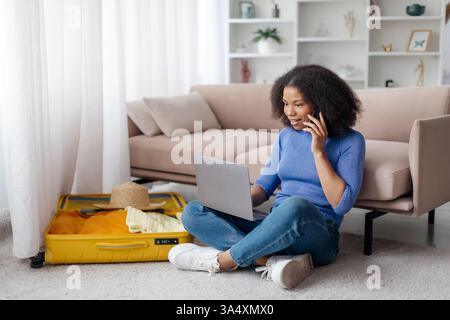 Happy young black woman with cellphone and laptop booking vacation from home Stock Photo