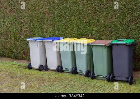 Different colored waste bins are neatly arranged along the side of a residential street. These containers include black, green, blue, and yellow bins Stock Photo