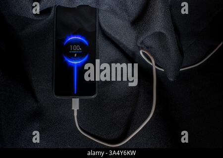 Mobile smartphone on a charger on a dark background. Battery indicators and charging process backlight on the screen. Stock Photo