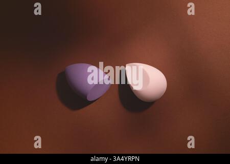 Cosmetics makeup sponges of different shape and color on a dark brown background with shadows. Stylish and artistic composition with beauty tools. Stock Photo