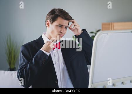 Young man in formal attire adjusting a red bowtie, looking thoughtful in a modern home interior with natural daylight and stylish decor Stock Photo