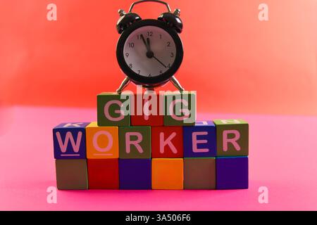 Colorful blocks spell out gig worker with an alarm clock on top, illustrating modern work trends in a bright setting. Stock Photo