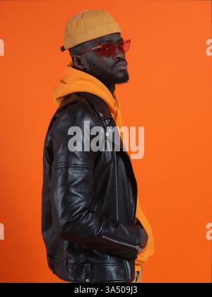 Side view of an African American man in a black leather jacket layered over a hoodie, wearing sunglasses and a cap against an orange background. A bold street style portrait suitable for fashion, urban lifestyle, and contemporary menswear campaigns. Great for urban fashion, casual wear, and accessory marketing. Stock Photo