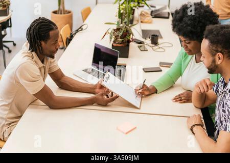 Three professionals collaborate in a modern coworking space, discussing ideas over a document. Ideal for business and workplace campaigns highlighting teamwork, diversity, and innovation. The high angle view from above captures an energetic, productive work environment. Stock Photo