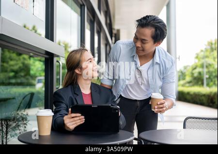 Thai business colleagues discuss work in a coffee shop, looking at each other as they talk. They appear professional and engaged in a collaborative meeting. A digital tablet is used to review documents, making this image suitable for business discussions and client meetings. Stock Photo