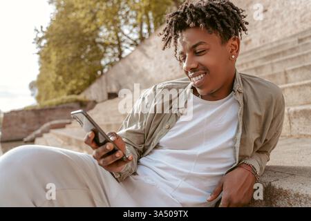 Smiling African American man with an Afro sits on outdoor steps by a river, using a smartphone in daytime. The casual moment highlights urban living, mobile technology, and relaxed street style. Great for topics on tech use, leisure, and contemporary fashion. Stock Photo
