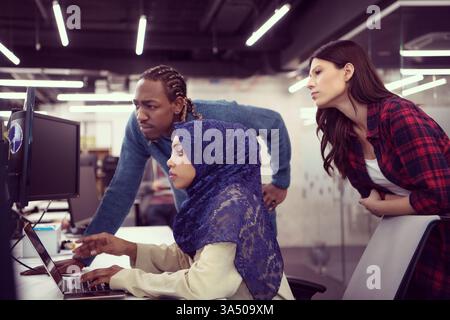 Black business people collaborating at a computer workstation with a white female coworker standing behind them in a modern office. This scene emphasizes teamwork, software development, and a diverse, inclusive workplace. Stock Photo