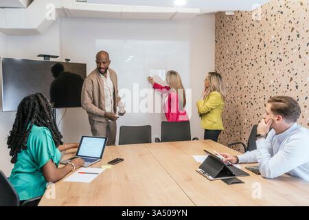Black businessman working with colleagues using laptop and writing on whiteboard while standing during meeting in office Stock Photo