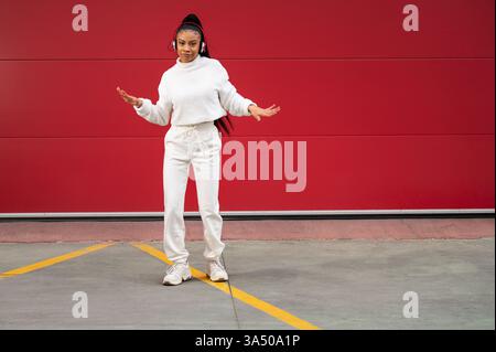 Energetic street dancer portrait of a smiling African American woman with long black hair wearing headphones, dancing in front of a vibrant red wall. This urban image is perfect for promoting hip hop, street style, and music lifestyle campaigns. Stock Photo