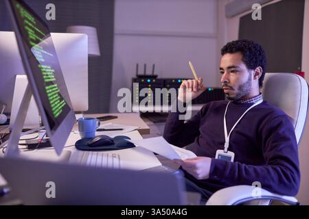 Side view portrait of an IT developer working at night, coding at a desk with documents. High-tech office setting conveys programming, cybersecurity, and software development themes for business use. Neon accents and bright screens add a contemporary feel. Stock Photo