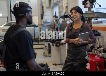 Two colleagues in a service garage discuss maintenance with a tablet, captured in daytime light. A diverse team collaborates in a busy workshop, highlighting intercultural teamwork and professional interactions. Stock Photo