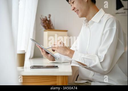 Cropped side view of a handsome young Asian man in a white shirt using a digital tablet in a coffee shop. The scene blends technology with a casual cafe setting, suitable for digital workplace and mobile tech themes. Great for business, freelancing and coworking content. Stock Photo