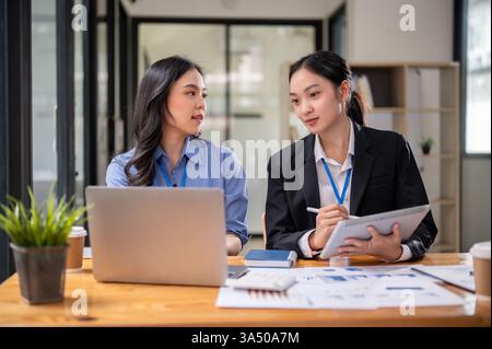 Two focused Asian businesswomen in a modern office discuss financial data and review reports together. They collaborate over laptops and paperwork, showcasing teamwork, planning, and professional finance and investment themes. This dynamic business scene is ideal for content about women in business and office collaboration. Stock Photo