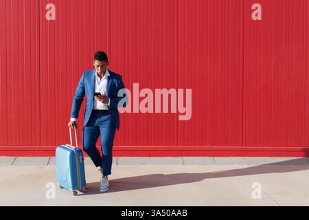 Hispanic man in a sharp business suit with luggage stands outdoors, messaging on a smartphone against a red wall. This image blends travel, work, and connectivity for modern professionals. Great for business, travel, and urban lifestyle contexts. Stock Photo