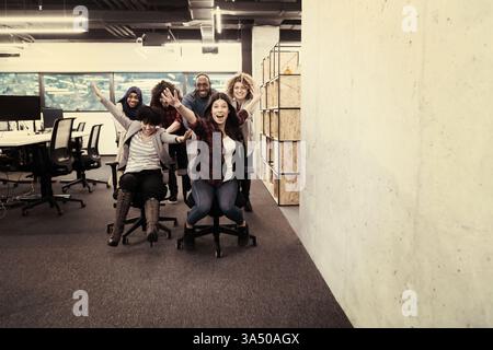 A dynamic and diverse startup software development team races office chairs in the workplace. The multiethnic group, including African American and White colleagues, share laughter and friendly competition. Stock Photo