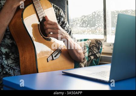 Crop shows a faceless male musician playing an acoustic guitar while watching a video lesson on a laptop in a van. A unique blend of music practice and digital learning for mobile or travel contexts. Great for content about online guitar lessons, remote musicians and education on the go. Stock Photo