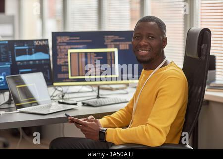 Portrait of a smiling Black man in a tech workplace, seated at a desk with code on computer screens in the background. A confident professional portrait for software development, IT, and cybersecurity themes. Suitable for business and technology campaigns. Stock Photo