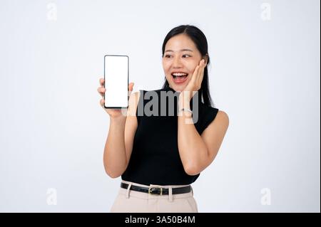 Asian woman shows a smartphone screen against a white background with a surprised expression. The clean, isolated composition makes this ideal for tech, app or online shopping campaigns. A versatile portrait for digital products and marketing materials. Stock Photo